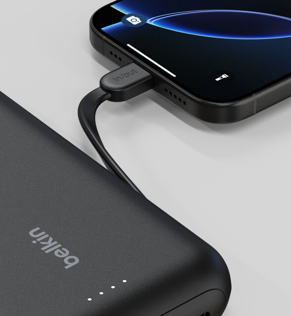 A close-up of a black Belkin BoostCharge Power Bank 20K with Integrated Cable connected to a smartphone, showing the "in/out" label on the cable and LED indicator lights on the power bank.