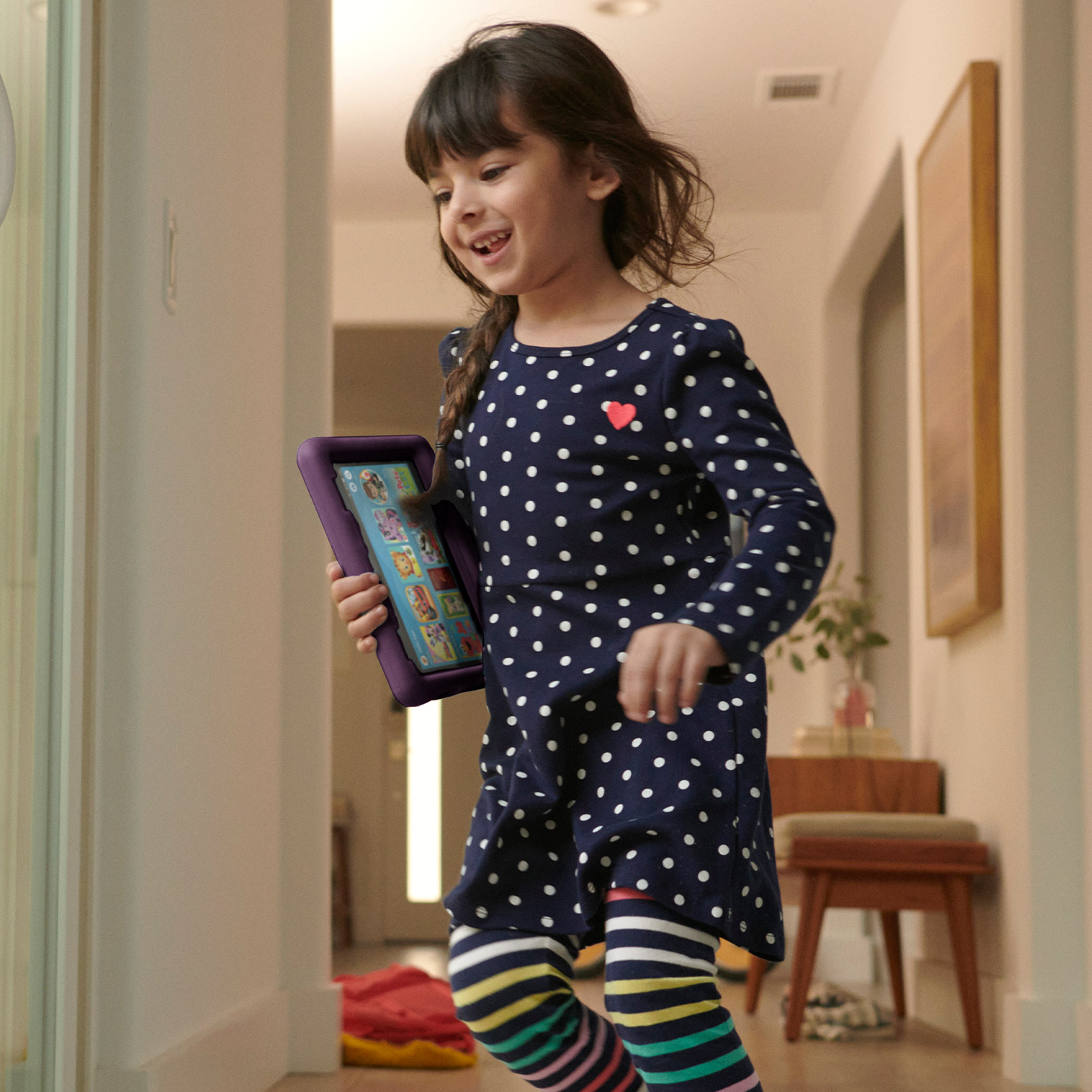 Bir yürümeye başlayan çocuk, mor kılıflı bir Fire HD 8 Kids tableti tutarak evin koridorunda koşarken gülümsüyor.