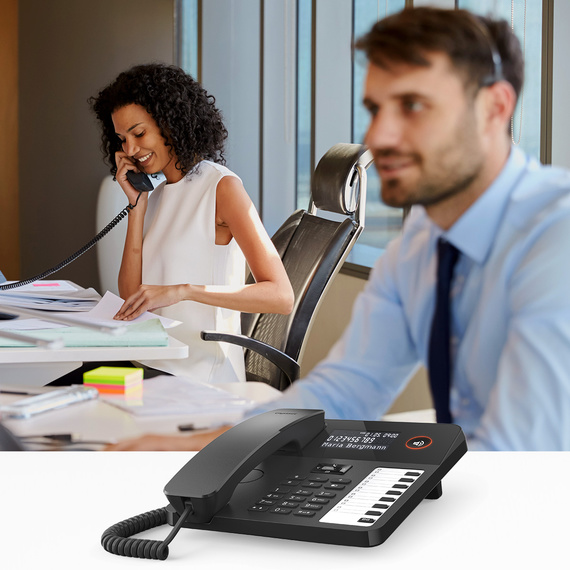 Ekranı ve fonksiyon tuşları olan modern bir masa telefonu, arka planda kulaklıklı bir adam ve telefonda konuşan bir kadın.