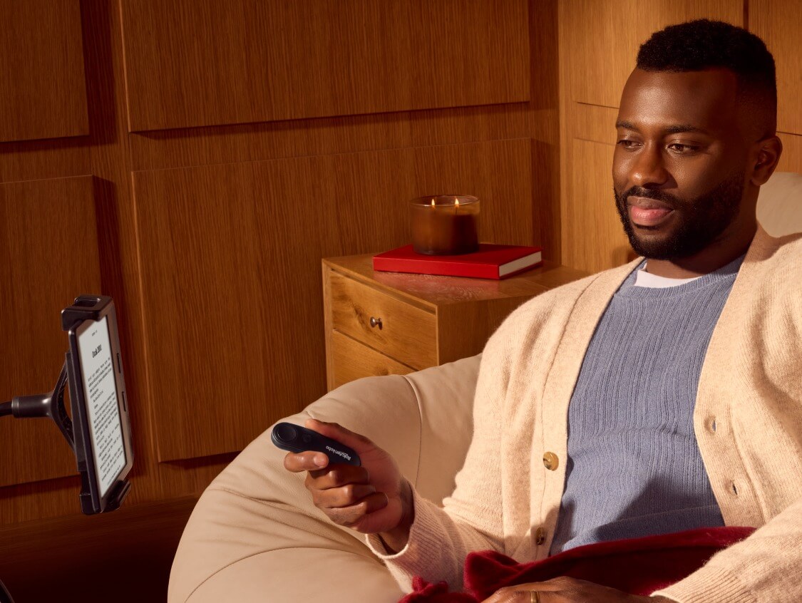 Rahatça oturan bir adam, sağ elinde Kobo Remote'u tutuyor ve bükülebilir bir stant üzerindeki Kobo e-kitap okuyucusuna bakıyor.