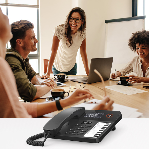 Ekranı ve fonksiyon tuşları olan modern bir masa telefonu, arka planda toplantıda bir grup.