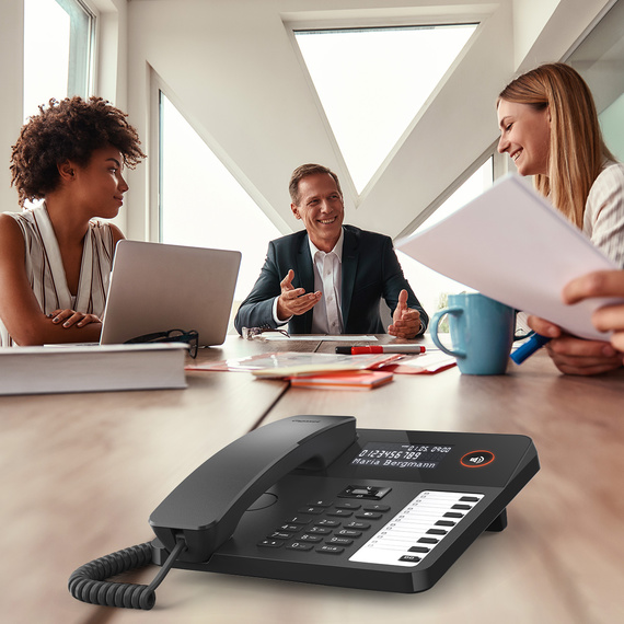 Ekranı ve fonksiyon tuşları olan modern bir masa telefonu, arka planda toplantıda bir grup.