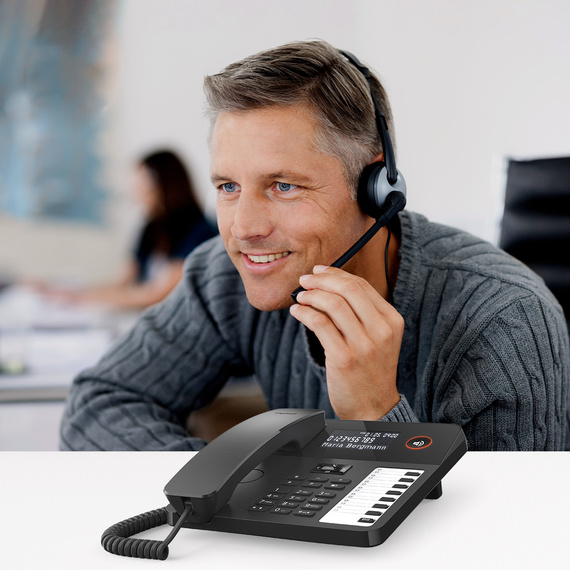 Ekranı ve fonksiyon tuşları olan modern bir masa telefonu, arka planda kulaklıklı bir adam.