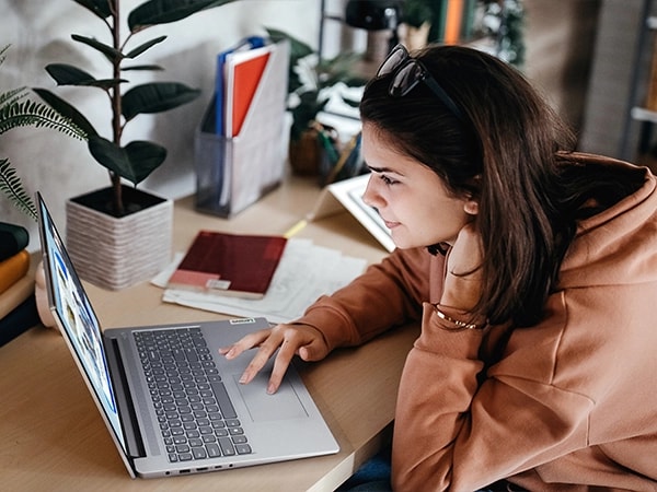 Çalışma masasına yerleştirilmiş Artic Grey renkli Lenovo IdeaPad Slim 3i Gen 9 16 inç dizüstü bilgisayar üzerinde çalışan bir öğrencinin yandan görünümü. Görüntü dizüstü bilgisayarın ve klavyesinin sol yan görünümüne odaklanıyor.