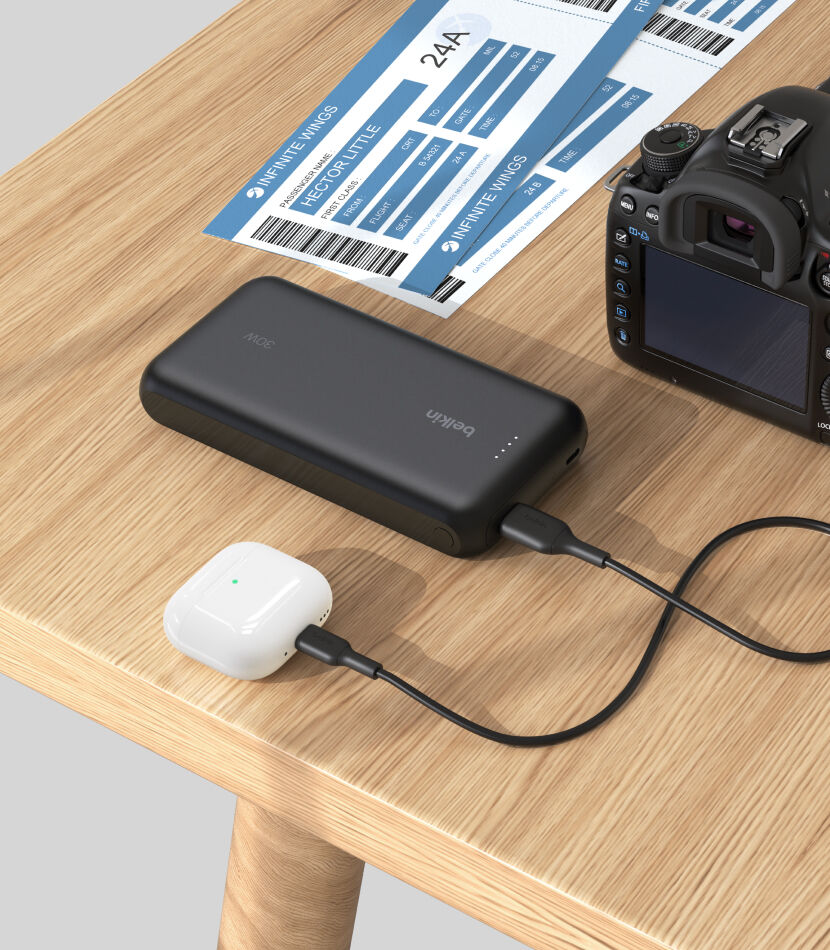 A black Belkin BoostCharge Power Bank 20K with Integrated Cable is placed on a wooden table, charging a pair of white AirPods, with a DSLR camera and boarding passes nearby.
