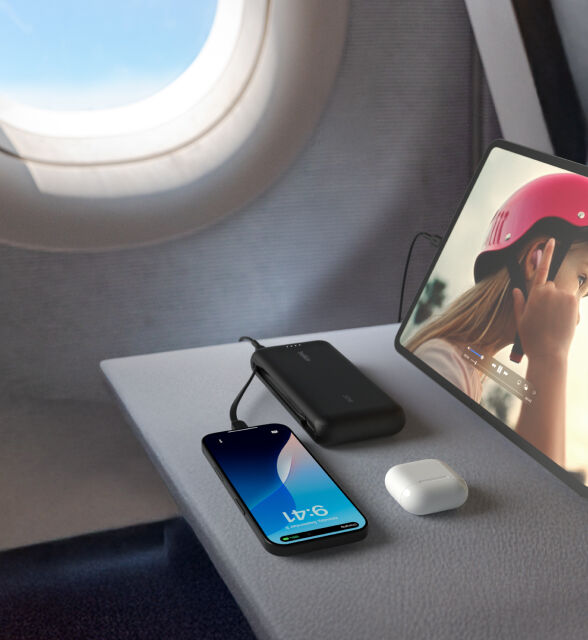 A black Belkin BoostCharge Power Bank 20K with Integrated Cable is placed on an airplane tray table, charging an iPhone and iPad, with a pair of AirPods nearby.