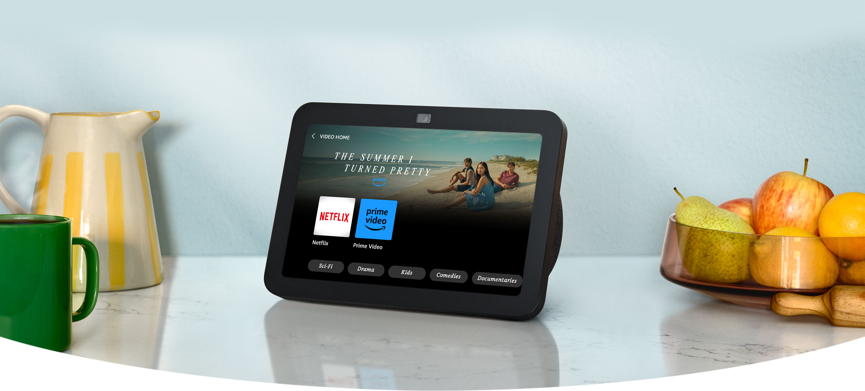 Echo Show 8, ekranda eğlence uygulamalarıyla birlikte mutfak tezgahında duruyor. Yakınlarda renkli bir sürahi ve meyve kasesi var.