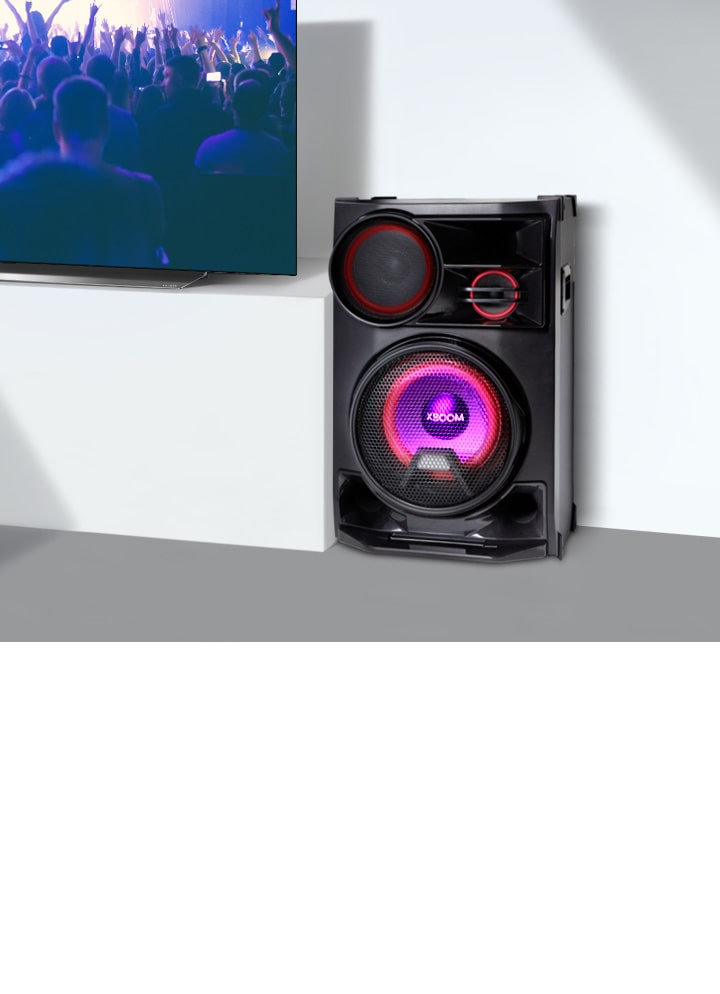 Görselde uzun beyaz bir TV masasının yakın çekim görüntüsü yer alıyor. Sol tarafta, bir LG TV konser sahnesini göstermekte. TV’nin sağ tarafında masaya bir hoparlör yerleştirilmiştir.