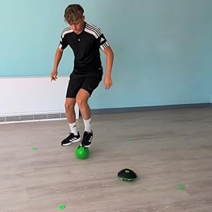 Siyah spor kıyafetleri giymiş bir kişi, spor salonunun zemininde küçük yeşil toplarla futbol becerileri çalışıyor.