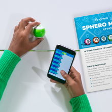 Basılı öğrenme kılavuzunun yanında blok kodlu Sphero Mini oyuncak robot ve telefon tutan çocuk.