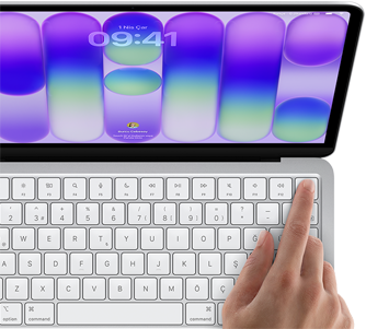 MacBook Neo, gümüş rengi, ekranda oturum açma penceresi gösteriliyor, kullanıcının parmağı klavyenin sağ üst köşesindeki Touch ID sensörüne dokunuyor
